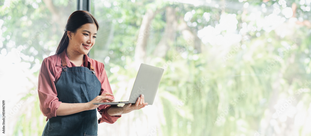 Poster a woman is smiling while using a laptop. she is wearing a pink apron and is standing in front of a w