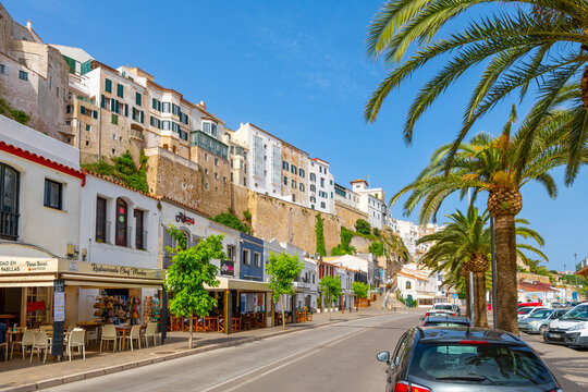 The whitewashed cliffside buildings with sidewalk cafes and shops below along the waterfront at the port harbor of Mahon, on the Mediterranean Balearic island of Menorca, Spain.