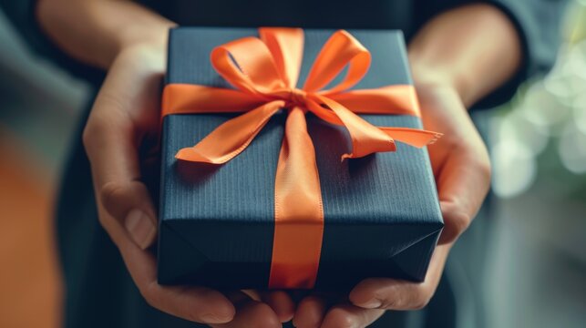 A close-up of hands holding a dark gift box wrapped with an elegant orange ribbon, symbolizing gift-giving and celebrations.