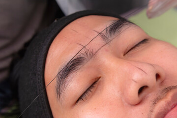 Using pre-inked threads to make guide lines around the eyebrows of a male patient to get te exact...