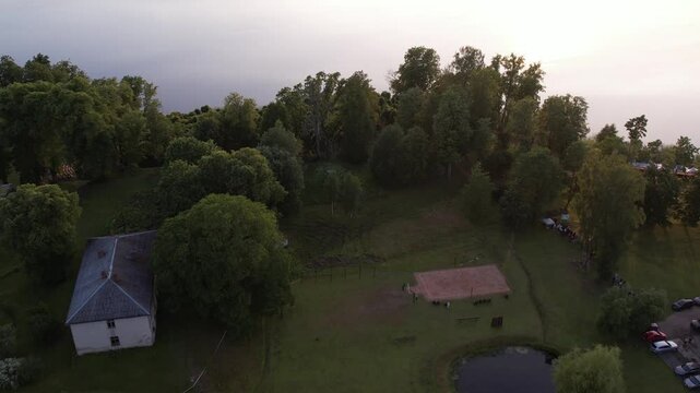 A stunning drone view of Katvari village at sunset, highlighting the Katvari linden tree by the lake. The golden sunlight casts a warm glow over the tranquil water.