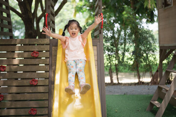 Asian little girl is sitting on a green slide at a playground. The playground is full of colorful...