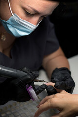 A nail technician, wearing black gloves, uses an electric nail drill on a client’s nail for precise detailing. 