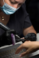A nail technician in black gloves uses an electric nail drill for detailed work on a client’s nail. The client’s hand is steady, highlighting the professional and precise manicure process.