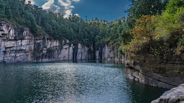 A beautiful emerald lake in the crater of an extinct volcano is surrounded by steep banks. Lush green vegetation on the rocks. Clouds in the blue sky. Madagascar. Tritriva Lake. - Powered by Adobe