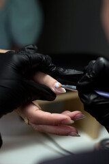 A nail technician, wearing black gloves, meticulously applies polish on a client’s nail with a thin brush. The client’s hand rests comfortably, showing the focus on precision and detail in the nail ca