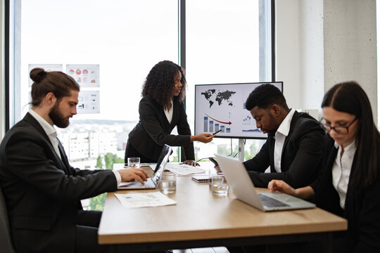 Diverse team in business meeting analyzing financial charts on laptop and screen. Professional environment with colleagues collaborating. Urban cityscape visible through large windows in background. - Powered by Adobe