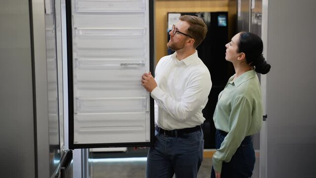 Salesman helping young woman choosing fridge in appliance store