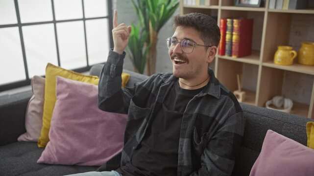 Young, happy hispanic man sporting glasses, seated on a sofa in a cozy home, points upward, brimming with a successful idea. number one smile on his face.
