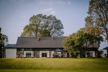 Blome, Latvia - September 11, 2023 - Large rustic house with stone and white walls on a grassy hill, surrounded by trees under a clear blue sky. Copy space.