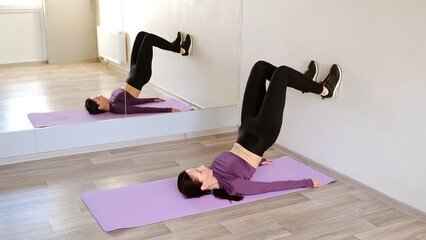 Athletic 30 year woman does the exercise in a lying position on the mat, lifting her feet off the wall and raising pelvis. Sporty girl pumps up the abs and legs muscles. Healthy lifestyle concept