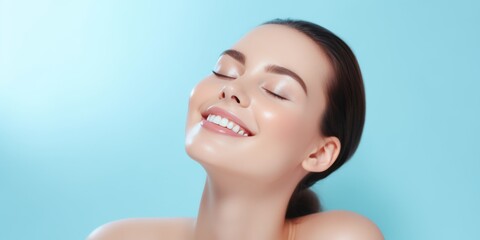 Young woman with radiant skin, smiling with closed eyes against a light blue background. She has a serene expression and glowing complexion