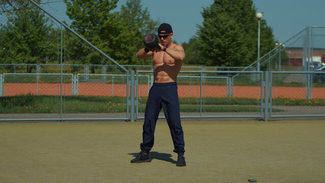 Young man training on outdoor sports field and performs kettlebell throw with both hands.Exercise to strengthen tendons, pumps up all muscles of arms and develops latissimus dorsi muscles