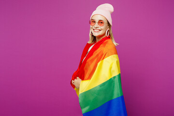 Side profile view young happy European lesbian woman wrapped in striped rainbow flag looking camera...