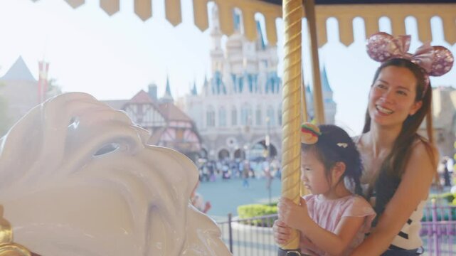 Asian mother and adorable kid daughter riding carousel at theme park.