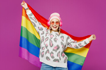 Obraz premium Young happy lesbian woman wear pink animal clothes listen to music in headphones hold rainbow striped flag isolated on plain purple background studio portrait. Pride day June month love LGBTQ concept.