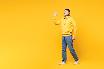 Full body side profile view young fun smiling cheerful happy man wearing hoody casual clothes walking going waving hand isolated on plain yellow orange background studio portrait. Lifestyle concept.