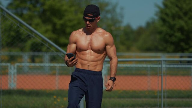 Young shirtless man showing off big biceps and abs while standing on an outdoor sports field with phone in his hand. Anaerobic training with equipment helps to work out and build all muscle groups