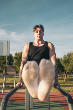 Calisthenic man doing L-sits on street workout bars. Focused young man with tattoos exercising on parallel bars at a park, doing abdominal sit-ups