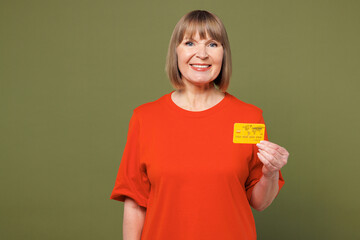 Elderly happy blonde woman 50s years old she wear orange t-shirt casual clothes hold in hand mock up of credit bank card isolated on plain pastel green background studio portrait. Lifestyle concept.