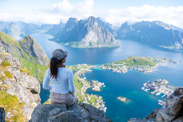 Naklejka premium A woman sits on a rocky cliff overlooking a stunning view of the Norwegian fjords, with mountains, clear waters, and a small town below. Reinebringen, Lofoten, Norway