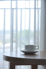 A white mug sits atop a wooden table next to a bright window, invitingly filled with tea drink. Concept Cozy Indoor Table.