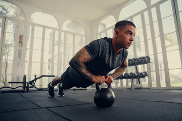 Young tattooed multiracial man in sportswear in plank position with kettlebell at gym