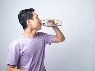 Young Asian man in a light purple shirt, drinking water from a transparent water bottle, standing sideways against a plain light grey background, highlighting hydration and healthy lifestyle.