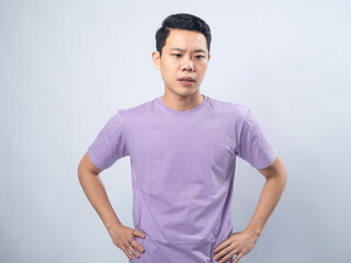 Obraz premium Young Asian man in a lavender t-shirt with a serious expression, standing with hands on hips. Studio shot on a plain background highlighting his casual outfit and confident stance
