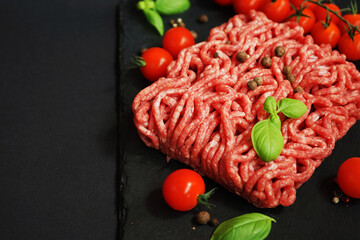 Fresh minced meat on a black slate tray next to cherry tomatoes, basil and spices on a dark...