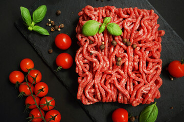 Fresh minced meat on a black slate tray next to cherry tomatoes, basil and spices on a dark...