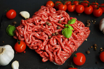 Fresh minced meat on a black slate tray next to garlic, cherry tomatoes, basil and spices on a dark...