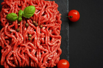 Fresh minced meat on a black slate tray next to cherry tomatoes, basil and spices on a dark...