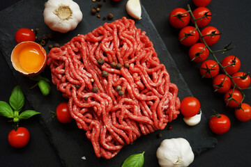 Fresh minced meat on a black slate tray next to garlic, cherry tomatoes, basil and spices on a dark...