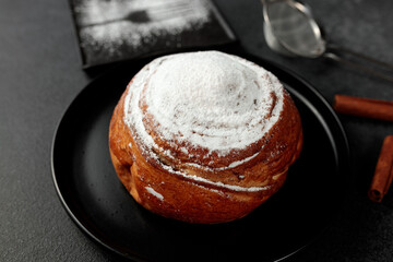Round, delicious bun with cinnamon and powdered sugar on a black plate on a gray background