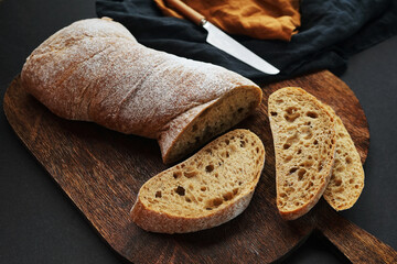 Fresh sliced ​​bread on a wooden board next to a knife with a linen napkin on a dark background