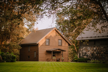 Obraz premium Blome, Latvia - September 11, 2023 - Charming wooden house with a small porch, surrounded by trees and greenery, adjacent to a stone building on a well-maintained lawn.