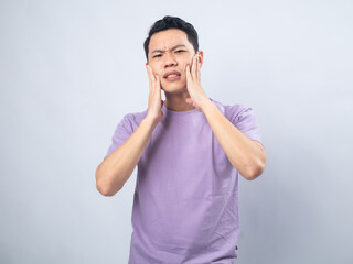 Fototapeta premium Young Asian man in a lavender t-shirt grimacing in pain while holding his cheek, indicating a toothache. Studio shot on a plain background, highlighting his discomfort and casual fashion.