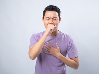 Young Asian man in lavender t-shirt coughing into his fist while holding his chest, indicating discomfort or illness. Studio shot on plain background capturing his casual outfit