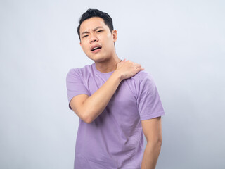 Fototapeta premium Young Asian man in a light purple shirt, grimacing in pain, holding his shoulder with one hand, suggesting shoulder pain or discomfort, standing against a plain light grey background.