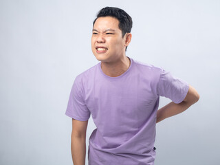 Young Asian man in a lavender t-shirt grimacing in pain and holding his lower back. Studio shot on a plain background, highlighting his discomfort and casual fashion.