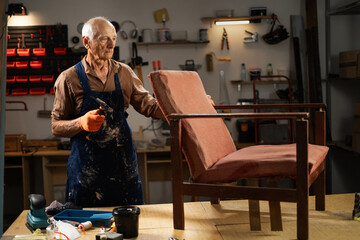 An elderly man in orange gloves and a blue apron begins to restore an old chair in a workshop. He...