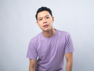 Young Asian man in a lavender t-shirt grimacing in pain and holding his lower back. Studio shot on a plain background, highlighting his discomfort and casual fashion.