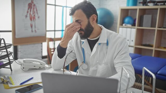 A middle-aged bearded man in a white lab coat working at a laptop in a clinic office, showing signs of fatigue or stress.