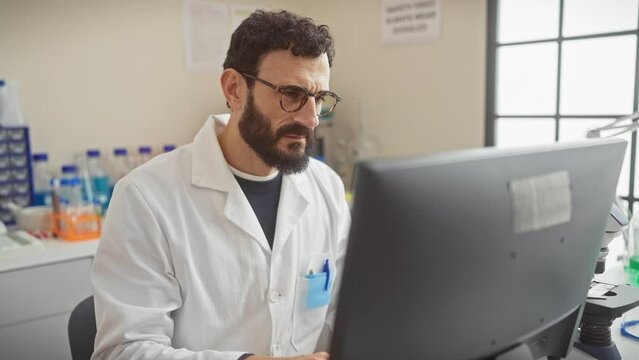 A thoughtful man with a beard works on a computer in a laboratory setting, showing concentration and expertise.