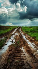 Fototapeta premium Muddy dirt road through green fields under a dramatic cloudy sky, stormy weather concept