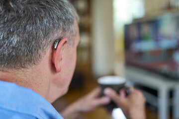 Close Up Of Mature Man Wearing Wireless Hearing Aid Watching TV At Home  Relaxing With Hot Drink