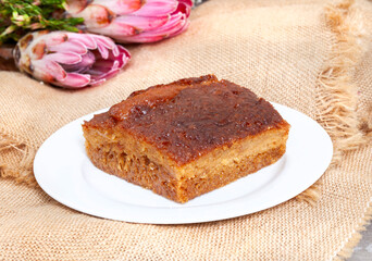 Traditional South African Malva pudding . one serving on a plate with burlap and copy space Protea flowers in background