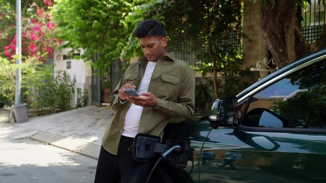 Medium long shot of busy Indian man with smartphone leaning on electric car charging with cable outdoors