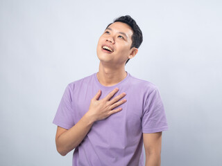 Young Asian man in a lavender t-shirt with a grateful expression, hands placed over his heart....